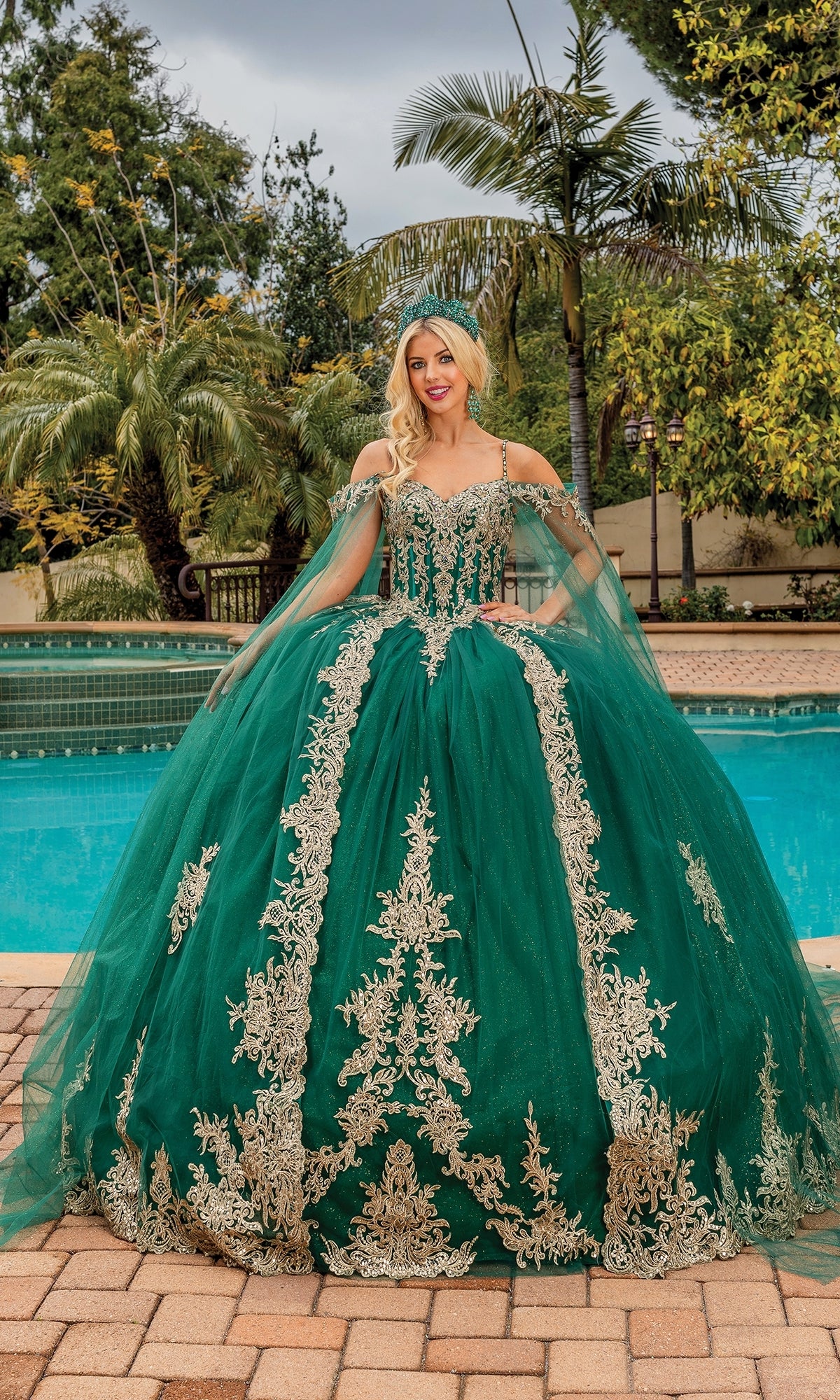 A person stands by a pool in the Quinceanera Dress 1768 By Dancing Queen, featuring gold embroidery and sheer cape sleeves, with palm trees and lush greenery as a stunning backdrop.